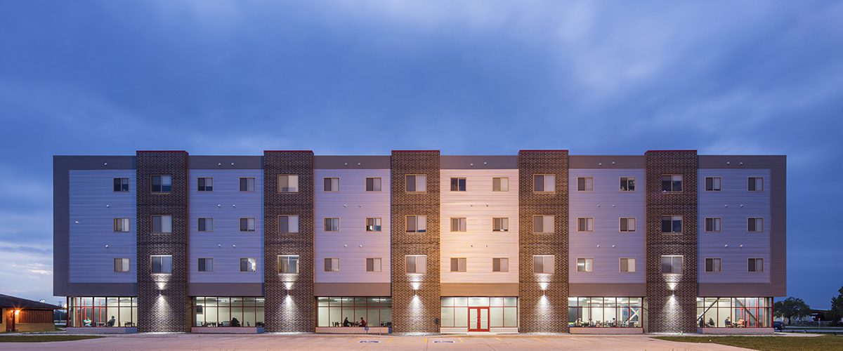 Exterior view of the SCC dorm building's full façade at dusk featuring architectural lighting with view into student lounge