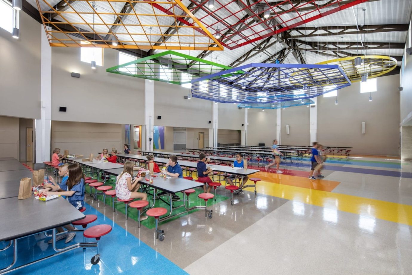 Elementary school cafeteria with children eating lunches. The floor is multi-colored and is mirrored by colorful, geometrical shapes suspended from the ceiling