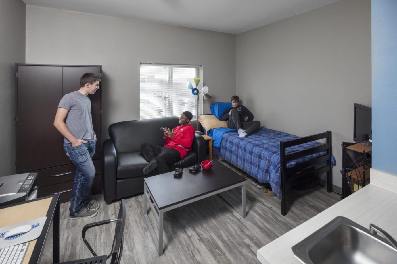 Dorm room featuring three students, a bed, couch, wardrobe, coffee table, desk, and sink