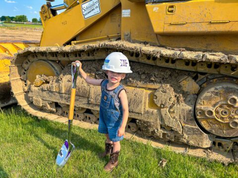 A little boy wearing a hardhat and holding a shovel in front of construction equipment